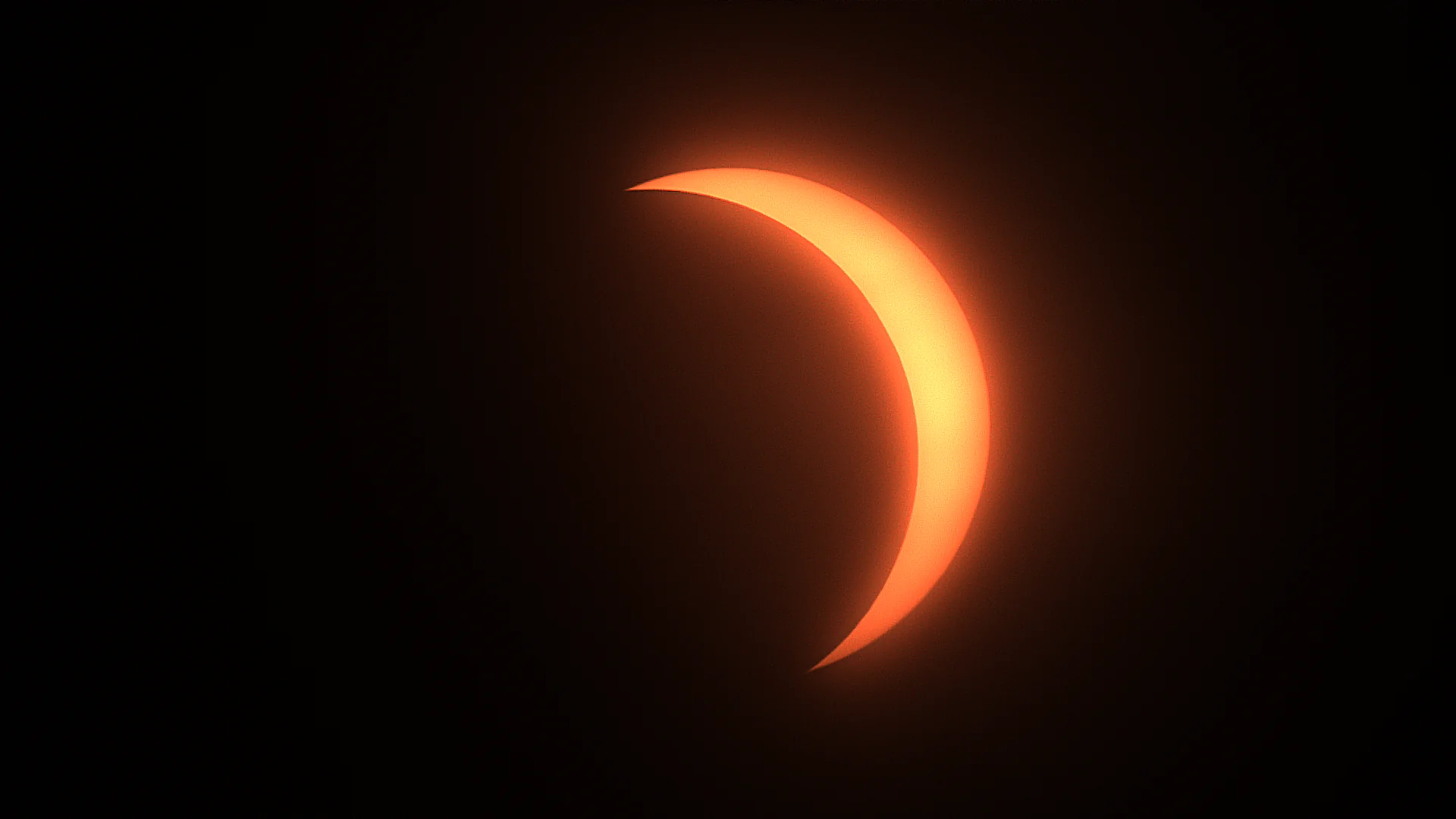 The partially eclipsed Sun photographed from Edinburgh. A thin crescent of glowing orange and pale gold light curves down the right of an otherwise black frame, its horns tapering to fine points top and bottom and its inner edge cut by the perfectly circular black silhouette of the Moon. A faint reddish glow surrounds the crescent. No sunspots or surrounding detail are visible.