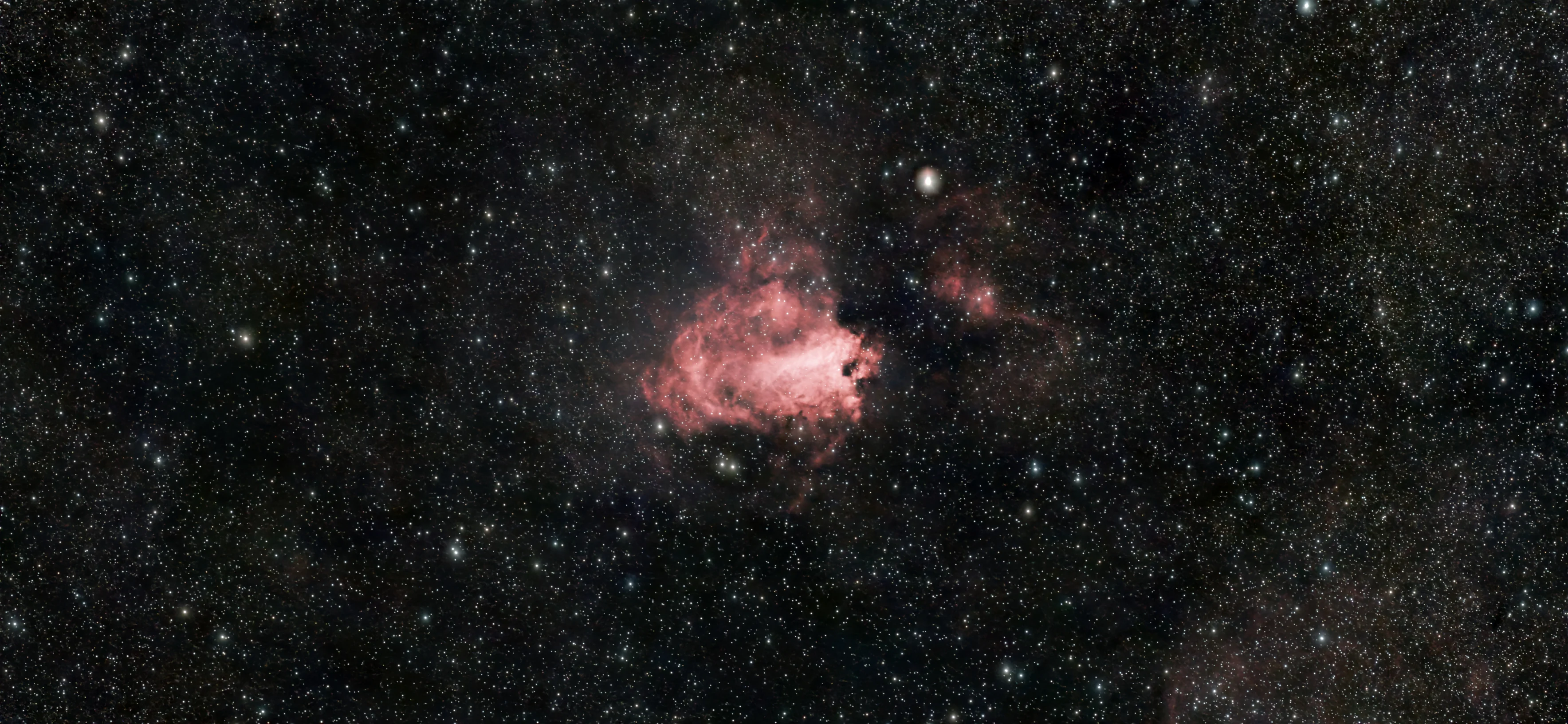 The Omega Nebula, M17, in a dense star field in Sagittarius. A small, bright salmon-pink cloud sits left of centre, its glowing core curling into a checkmark-like hook shape with fainter wisps trailing off it. A prominent white star sits just above and to the right of the nebula, with dim rust-coloured nebulosity spreading further right across the frame. Thousands of faint foreground stars crowd every part of the image, far denser than a typical field, marking the direction of the Galactic centre.