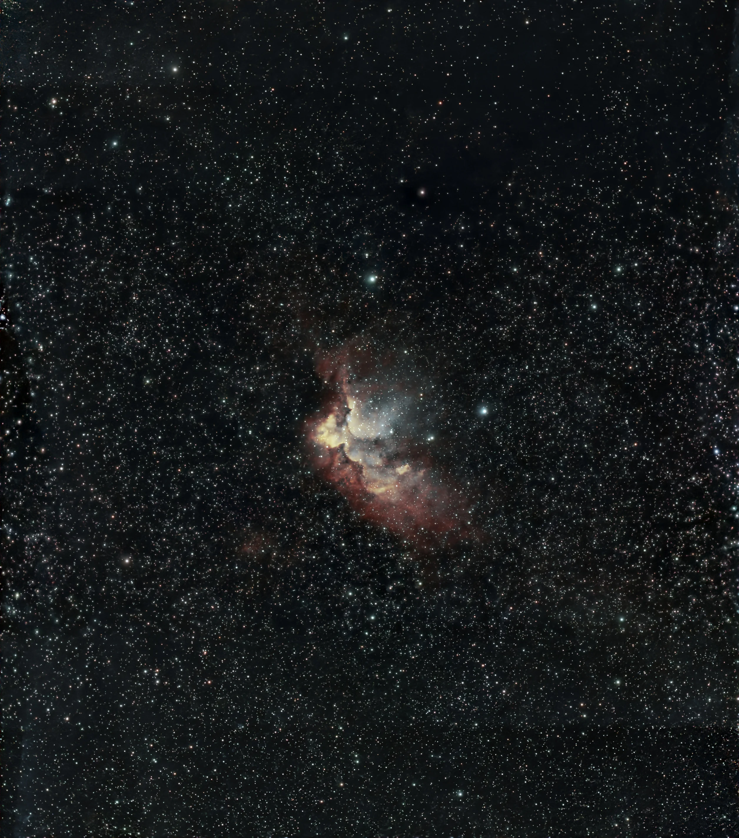 The Wizard Nebula, NGC 7380, near the middle of a dense Cepheus star field. A bright pale blue-white cavity holds the young cluster's stars, wrapped in salmon-red hydrogen, with cream-yellow ridges of gas along its lower left edge. A dark dust lane runs down through the centre of the nebula, a bright blue-white star sits just to its right, and thousands of white and pale gold stars fill the rest of the tall frame.